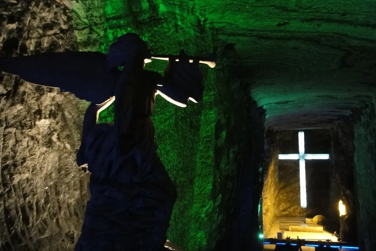 An angel statue blowing a trumpet in silhouette against green-lit salt walls, with a glowing white cross at the end of the tunnel