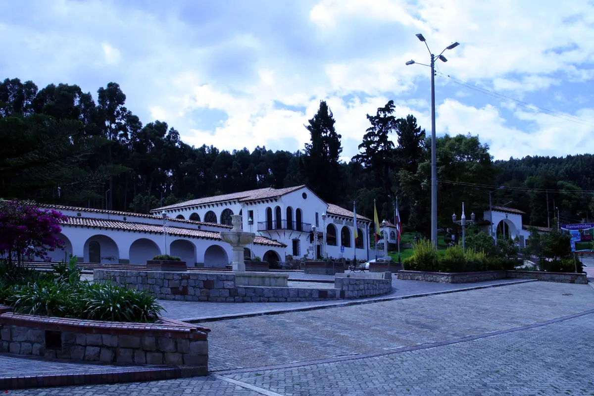 A white arcaded building with a fountain in a paved plaza at the Parque de la Sal, with eucalyptus forest behind