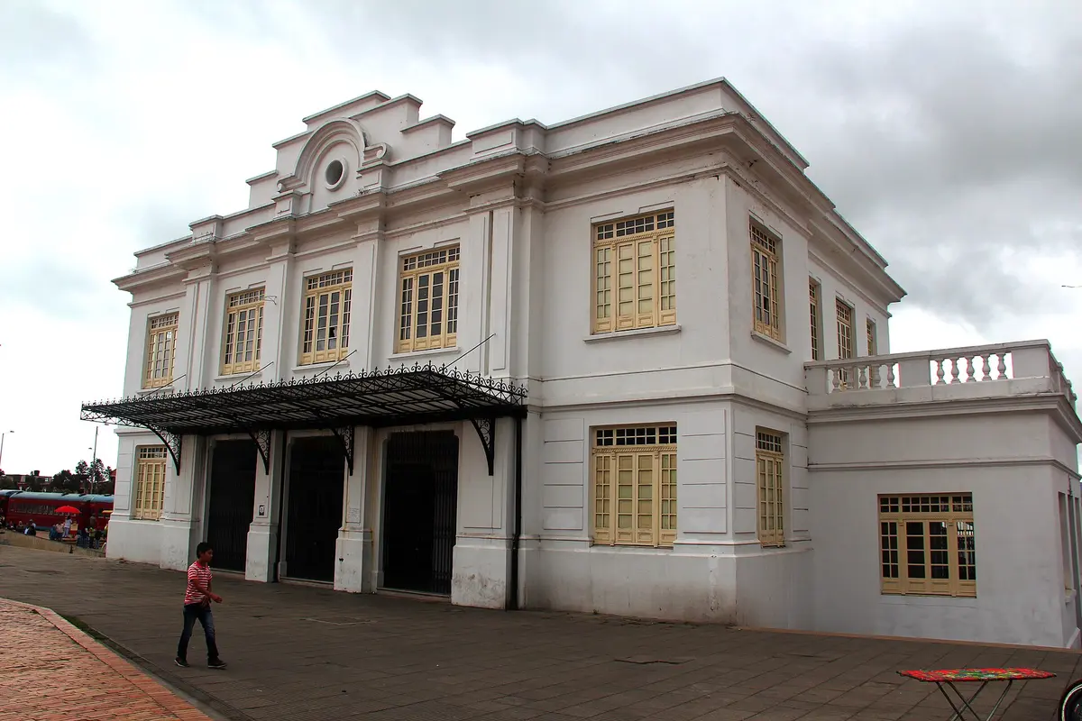 A two-storey white railway station building with a black iron canopy over its arched doorways, and a red train carriage at the far left
